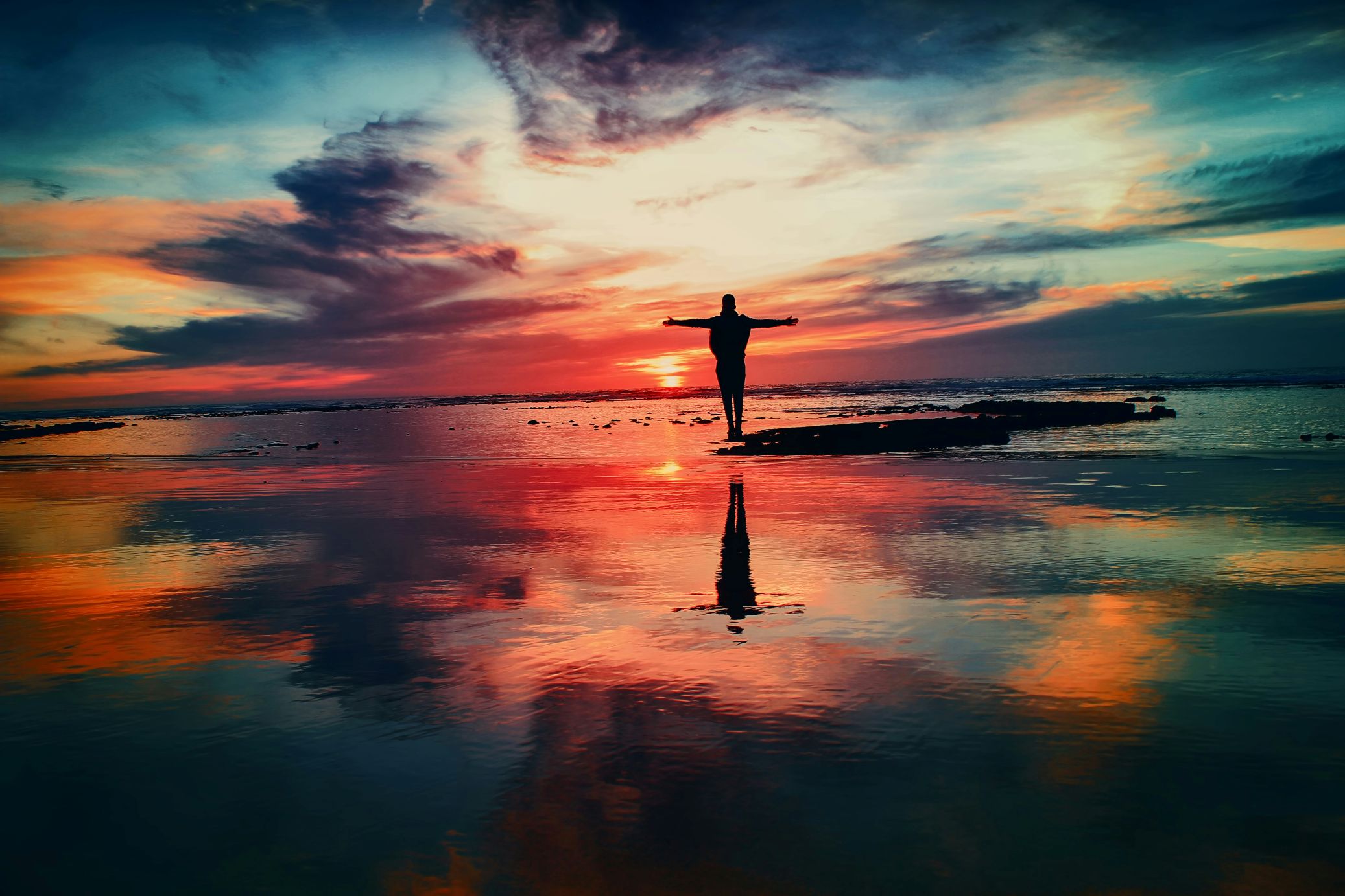 Silhouette of a person with arms outstretched on a beach at sunset, with their reflection in the wet sand.