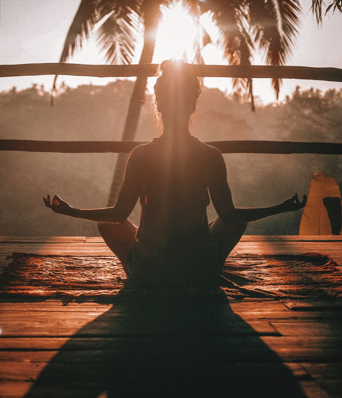 Silhouette of a person meditating on a wooden deck at sunrise, with palm trees in the background.